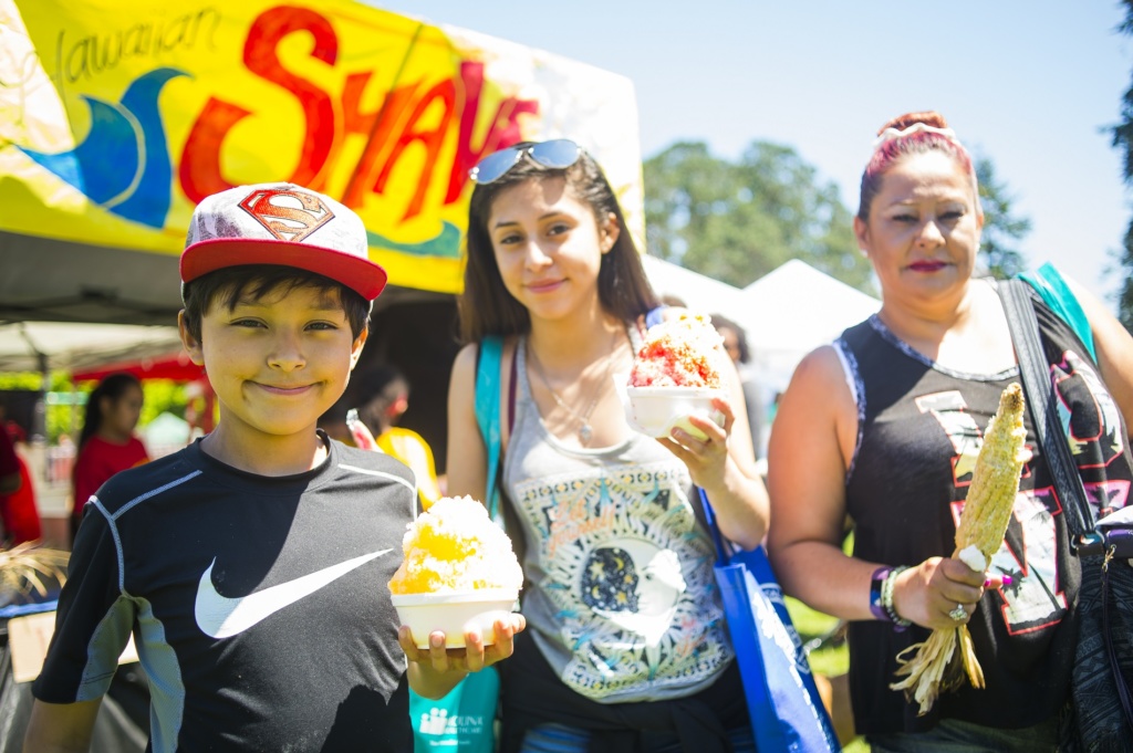 Three people stand facing the camera holding festival food in front of them - shave ice for the kids and roasted corn for the adult - at an outdoor festival with brightly colored booths behind them.