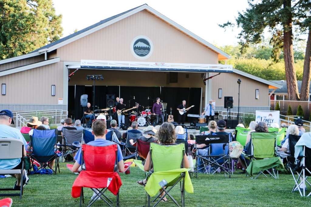 Lakewood, WA Pavilion in the Park on a summer evening at sunset with a crowd of people sitting on brightly colored folding chairs on the grass listening The Lonely Hearts Club perform on stage in the background of the image. In the foreground a man and a woman sit in a bright red and bright green chair.