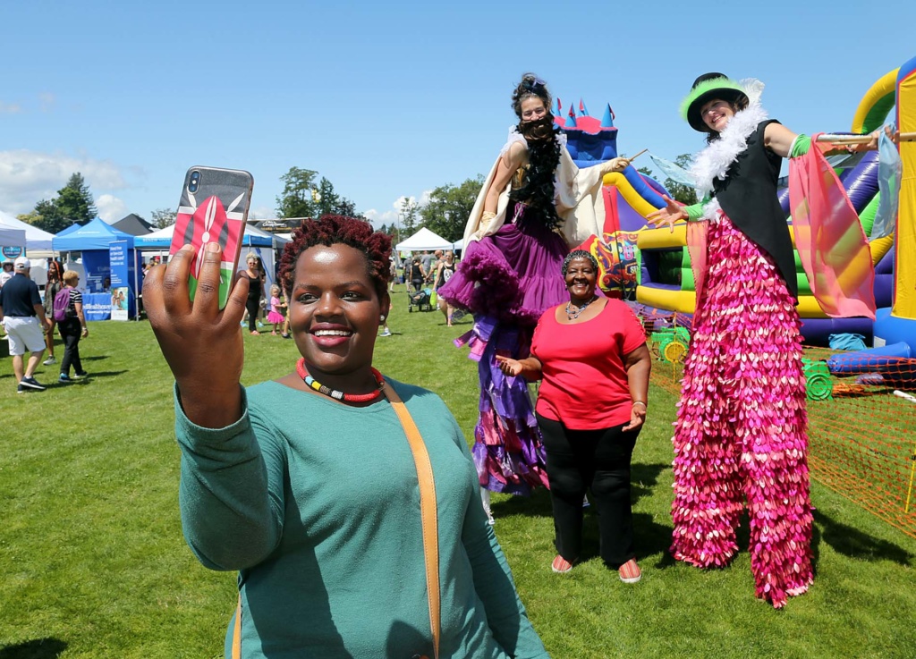 A person holds up a cell phone to take a selfie with a friend in the background who is standing next to two people dressed in costumes on stilts. Behind them is the setting of SummerFEST, an outdoor festival with booths and play equipment.