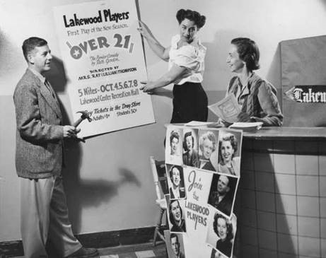 Black and white photo with a man and two women at the Lakewood Players Theater, posed to show them hanging up a poster about an upcoming show. A historic image from Lakewood, WA