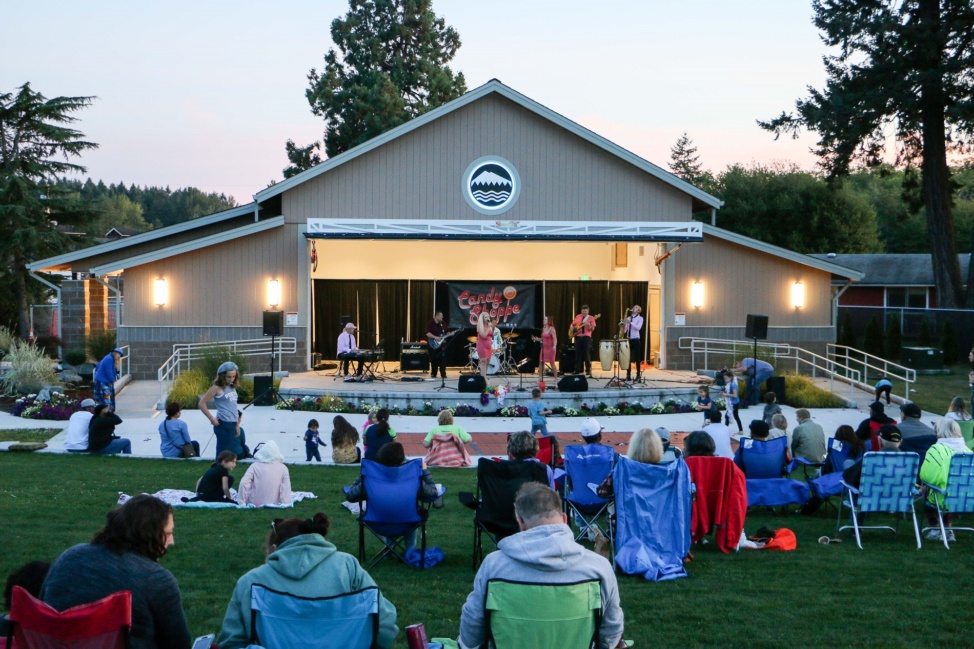 Pavilion at Fort Steilacoom Park in Lakewood, WA at sunset with a band performing on stage and a crowd seated in multi-color folding chairs on the grass enjoying the show.
