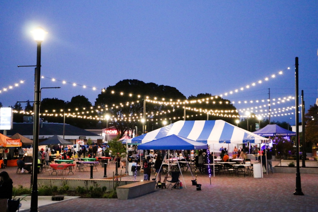 Blue and white canopy set up over tables at the Colonial Plaza grand opening event in Lakewood WA with white lights strung from poles and a darkening sky.