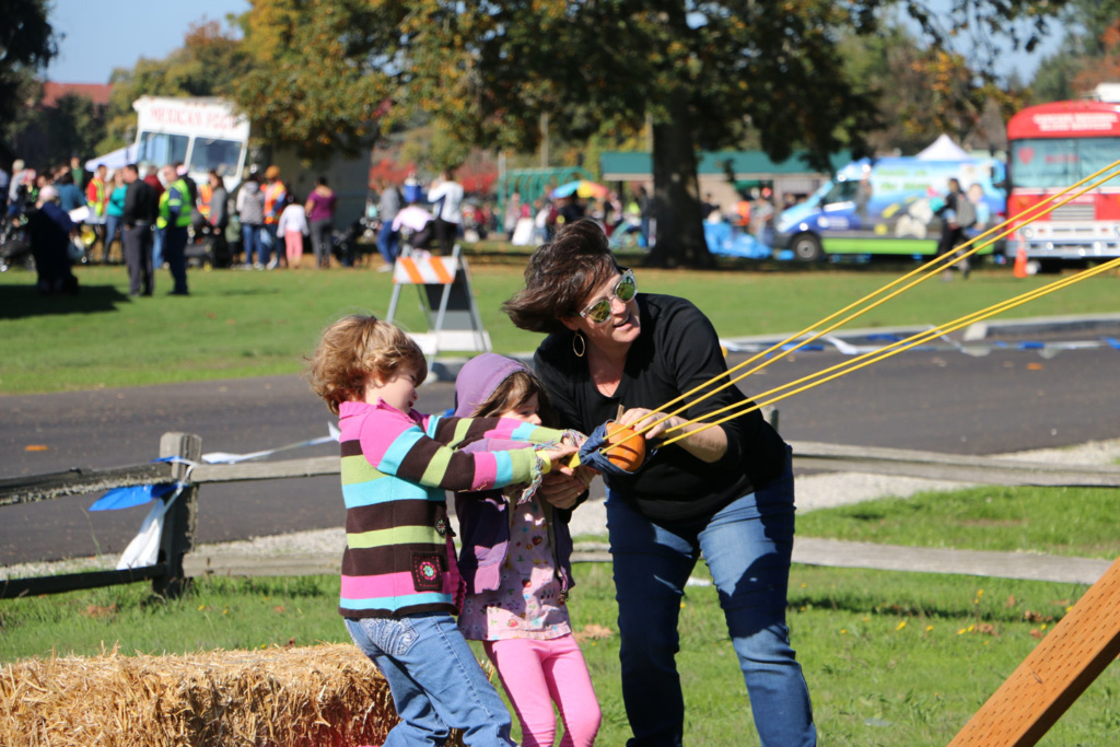 A mom helping two little girls pull back a slingshot loaded with a pumpkin.