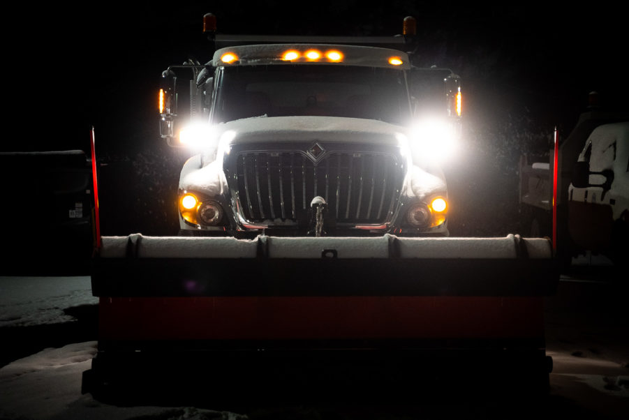 A truck with snowplow on front in the dark and headlights lit