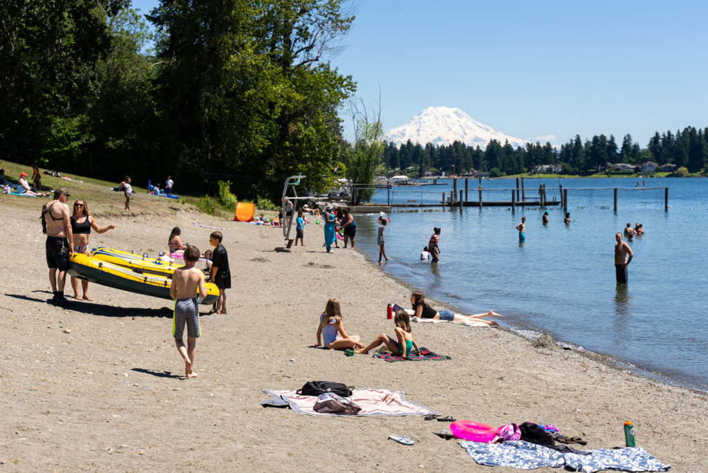 The beach at American Lake park on a sunny day. There are lots of people playing in the water and sand and Mt. Rainier is in the background. 