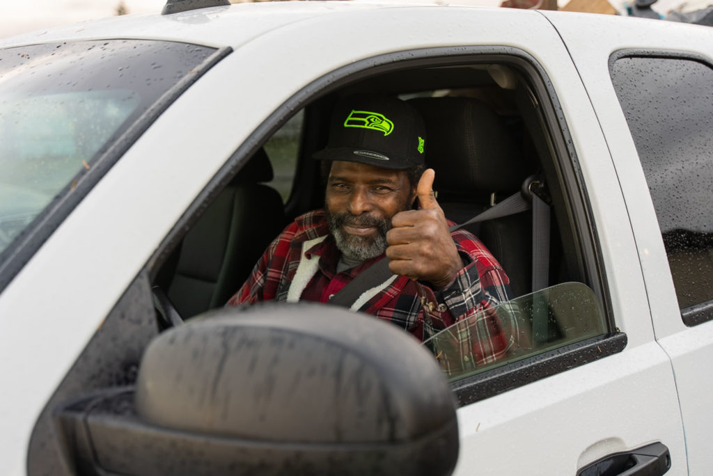 A man sits in the driver's seat of a white vehicle and gives a thumbs up to the camera