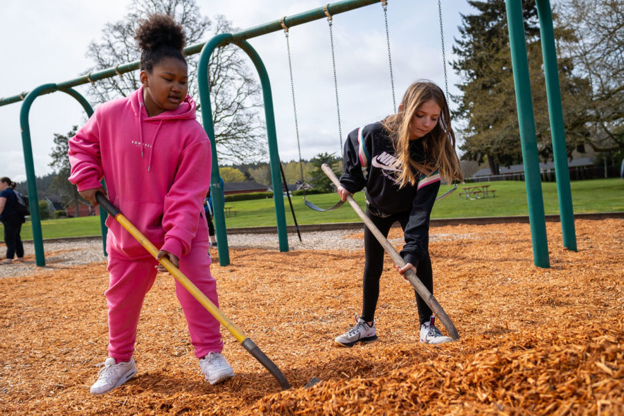 Two young people use shovels to spread woodchips at a playground during a volunteer event.