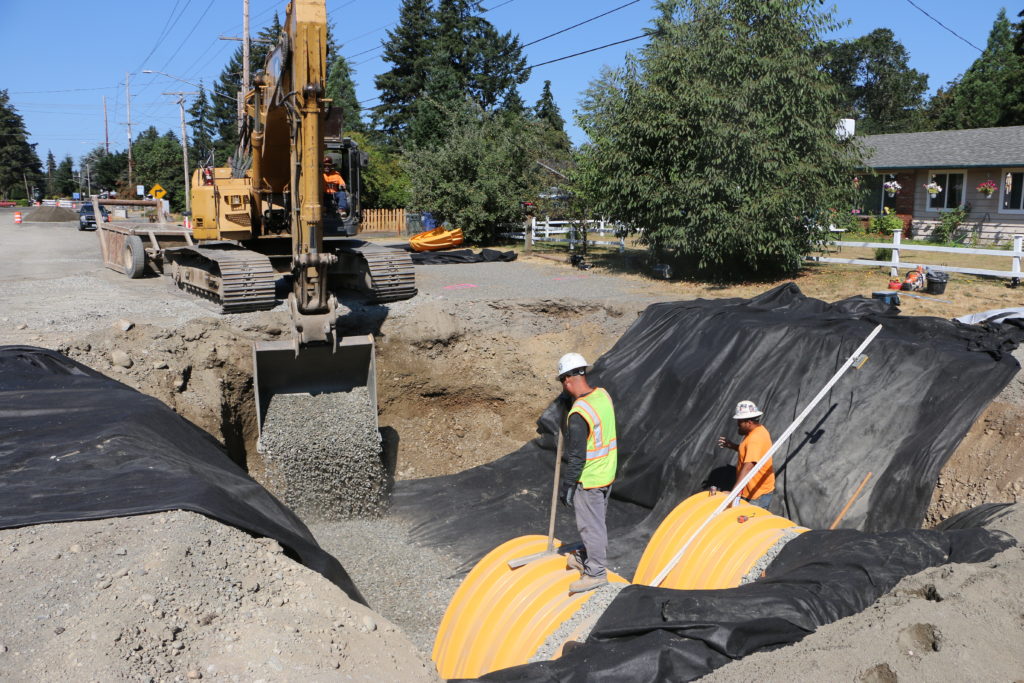 Construction to install major stormwater infrastructure in Lakewood with a digger dropping gravel in a large hole and two men prepared to move it around.