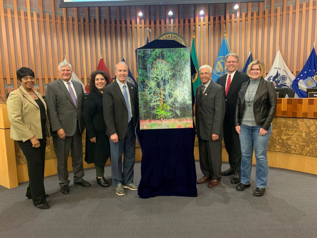 Members of the Lakewood City Council stand in front of Council Chambers around a painting of an Oak tree on loan by a local artist.