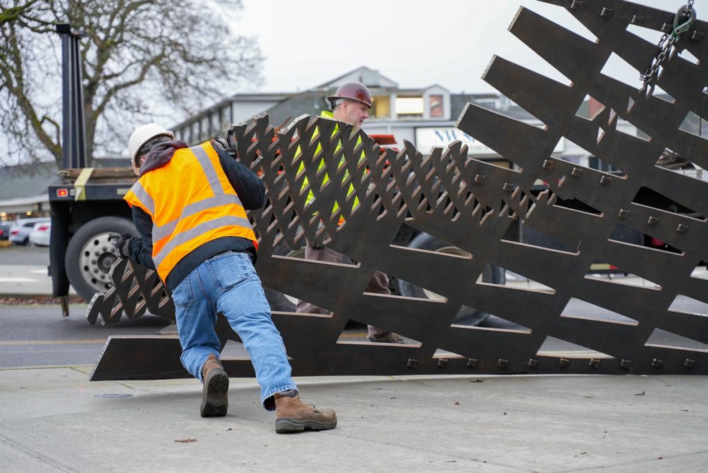 A worker in a hard hat and orange vest leans into a large metal piece of public art at Motor Avenue in Lakewood, WA where the piece is being installed.