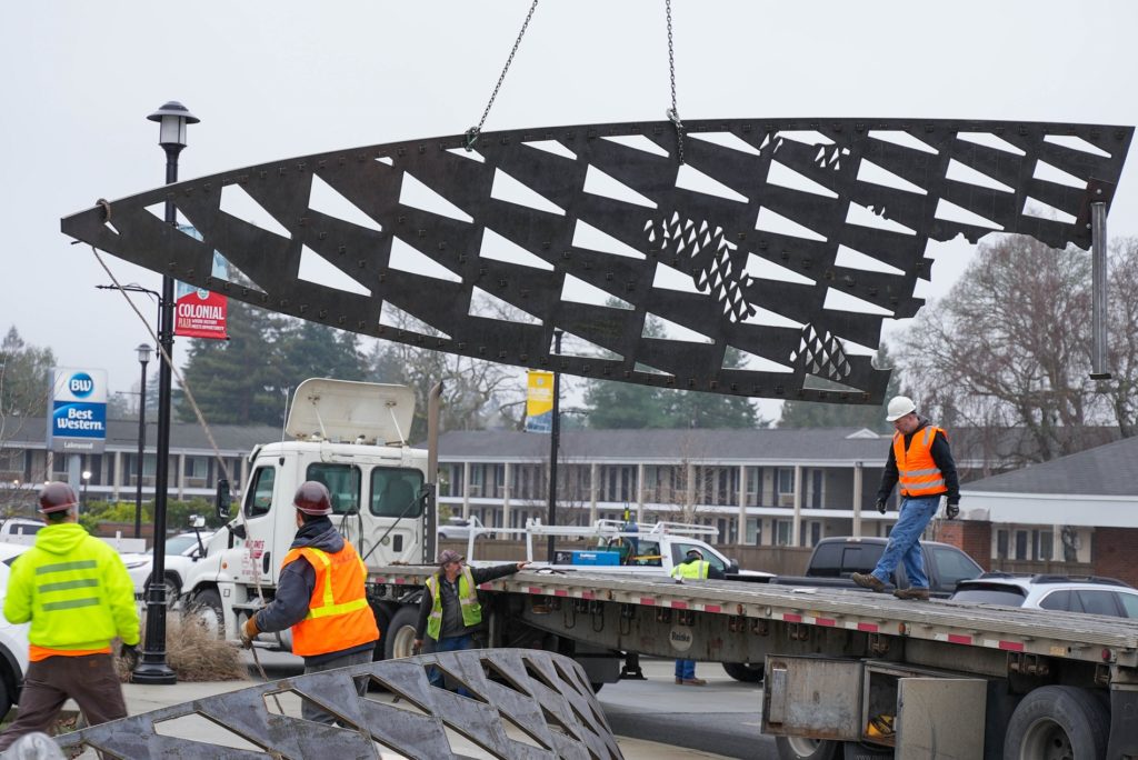 A triangular piece of metal public art is lowered from a trailer bed by a large crane to the ground in Lakewood, WA.