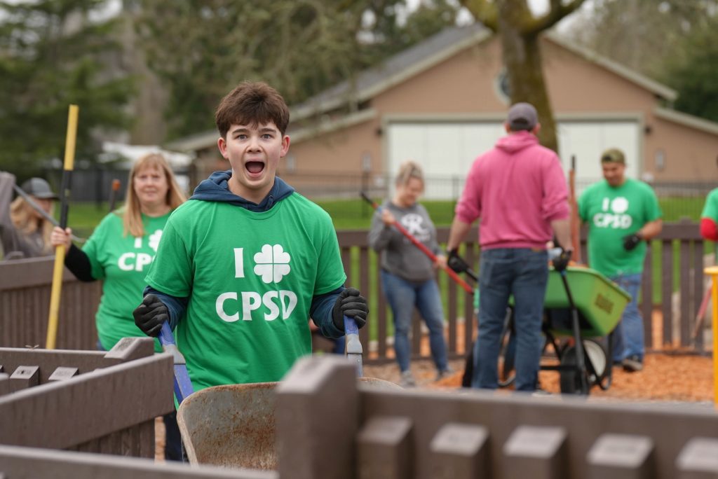 A student in a green t-shirt makes a funny face at the camera while wheeling a wheel barrow.