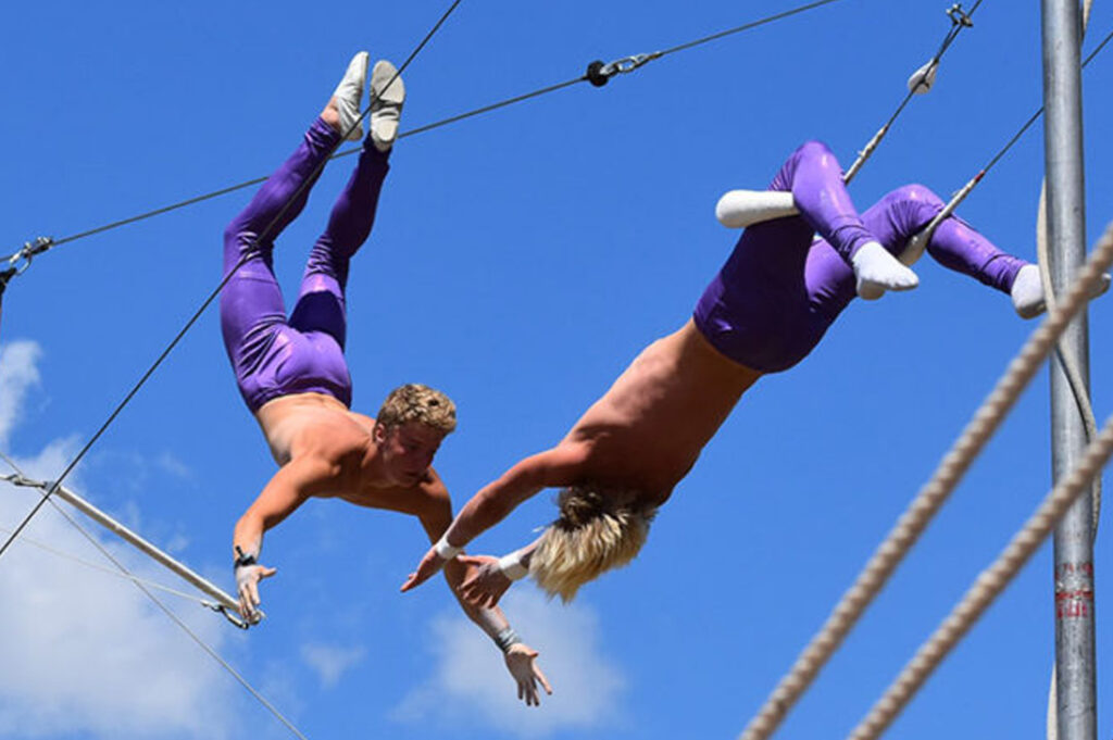 Two male trapeze gymnasts wearing purple pants perform tricks.