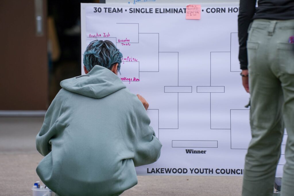 A member of the Lakewood Youth Council crouches and writes the results of their corn hole game on the tournament bracket. 
