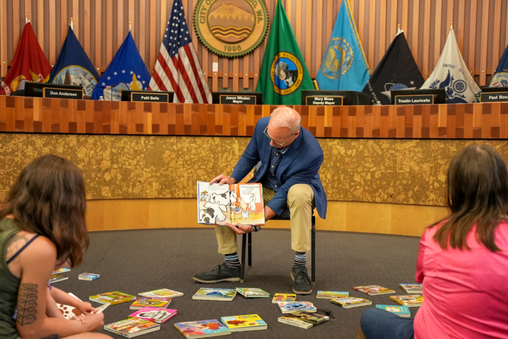 Thị trưởng Lakewood Jason Whalen cầm một cuốn sách dành cho trẻ em trong khi ông đọc cho hai bà mẹ và con cái của họ nghe tại sự kiện Kể chuyện của Thư viện Quận Pierce tại Tòa thị chính Lakewood trong Phòng Hội đồng vào ngày 1 tháng 2023 năm XNUMX.