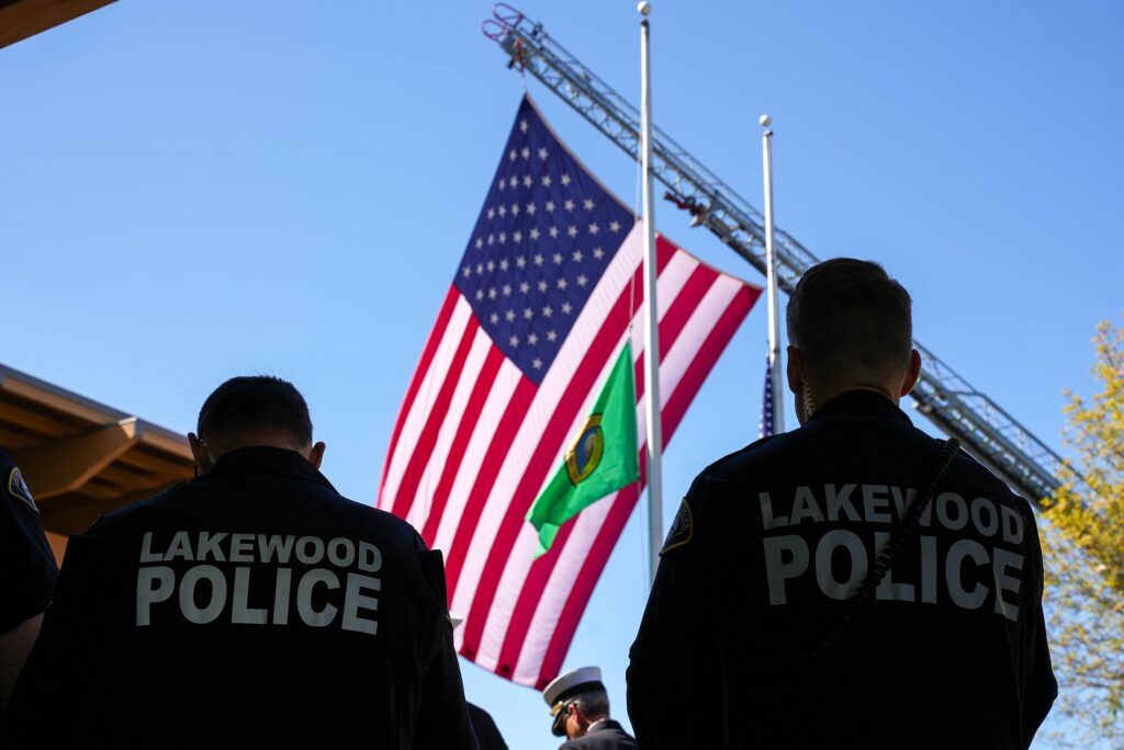 Le drapeau américain flotte depuis une échelle de camion de pompiers au-dessus de l'hôtel de ville de Lakewood. Au premier plan, deux policiers de Lakewood se détachent sur le ciel bleu.