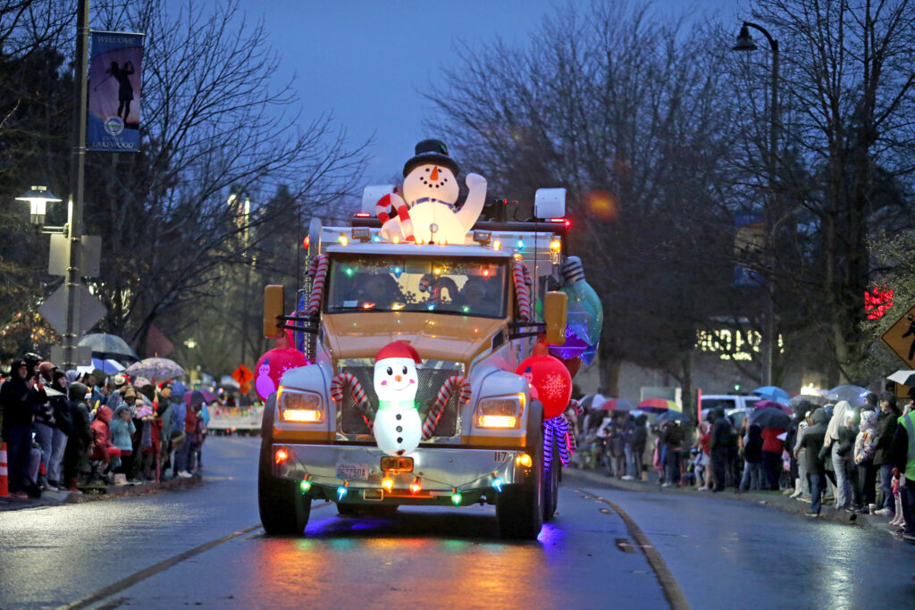 A large truck with christmas lights and snowmen on it drives up the street. People are gathered on both sides of the street watching the parade.