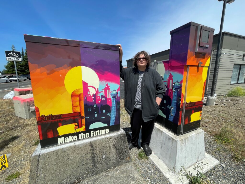 Lakewood high school student Sam Reed poses next to the two signal boxes that hold his artistic design in Lakewood at the corner of 96th Street SW and South Tacoma Way SW.