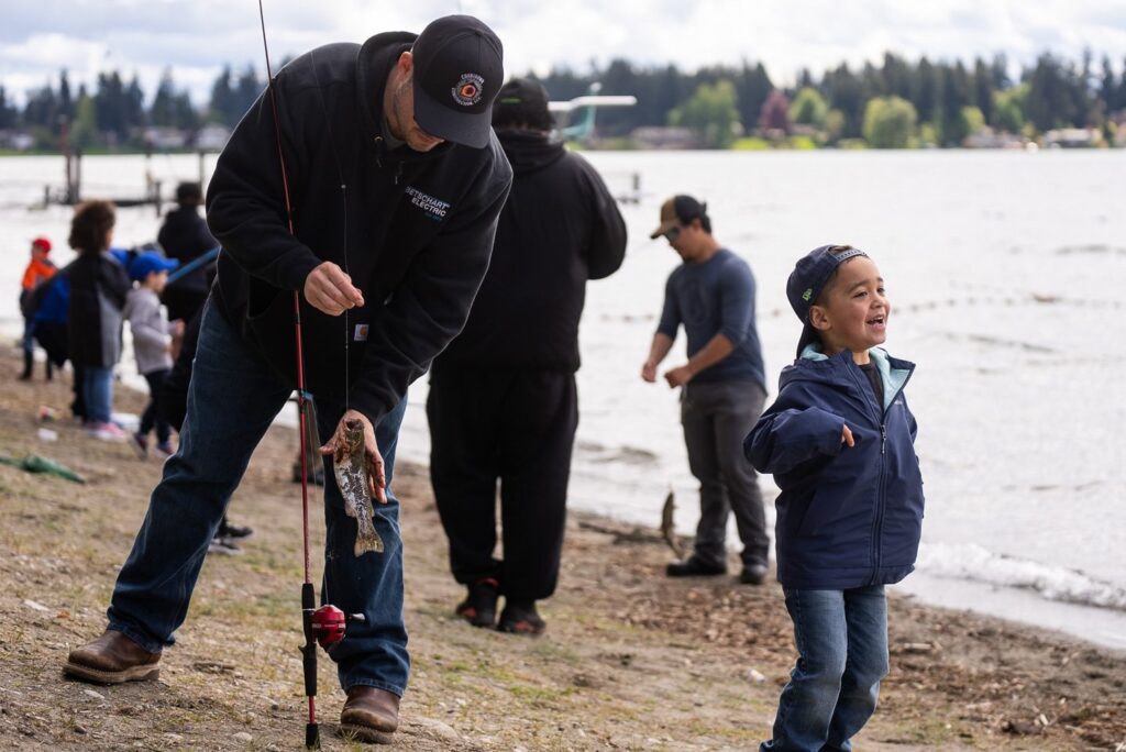 A man tries to grab a fish hanging off of a fishing line while his toddler son smiles widely and laughs.