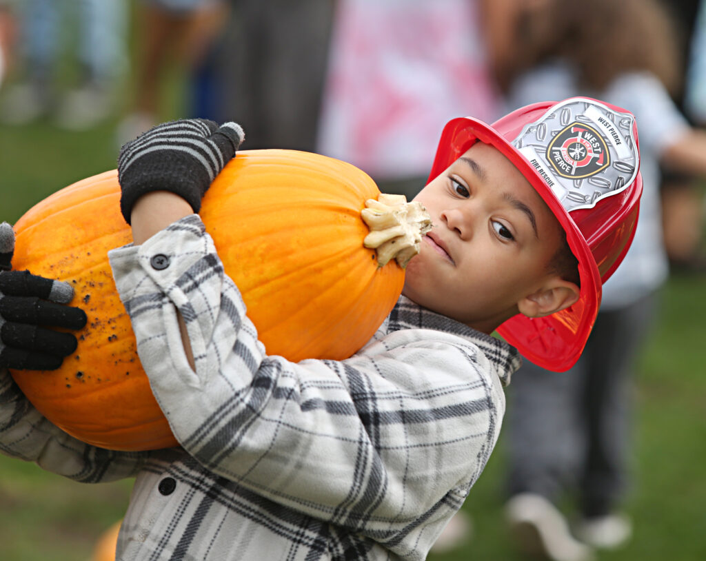 A child leans back as he holds a large pumpkin at the Lakewood Truck & Tractor Day 2023 event. He is wearing a red West Pierce Fire & Rescue plastic hat.