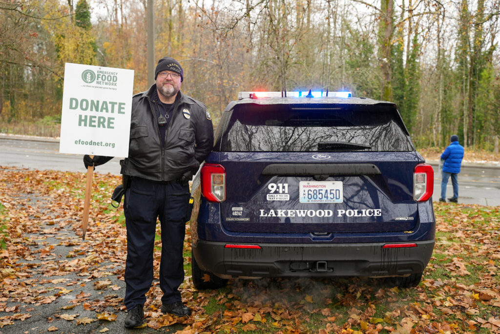 Lakewood Police Lieutenant holds a sign that says "Donate Here" while standing next to a squad car.