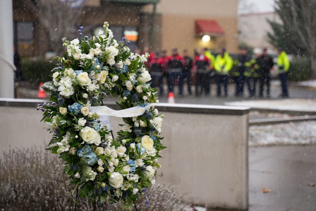 A wreath with white and blue flowers stands in the foreground with people blurred in the background at the 2022 Lakewood Police flag raising ceremony.