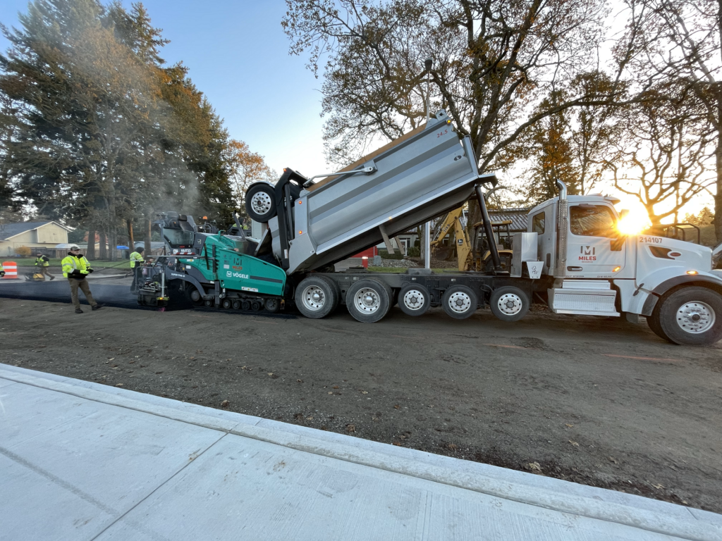 A dumb truck tilts its bed back while the sun shines through the trees. The truck is paving Washington Boulevard in Lakewood on Nov. 8, 2023.