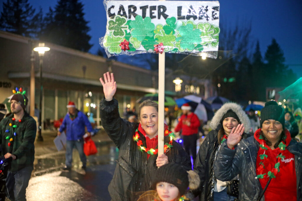 A person holds a sign that reads Carter Lake and waves at the camera with a smile in the 2023 Lakewood Holiday Parade of Lights