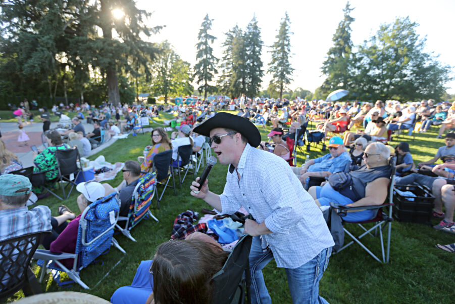 A singer in a cowboy hat and white button up shirt and jeans leans into a handheld microphone while walking into a seated crowd in an outdoor concert venue with trees and grass and people in folding chairs.