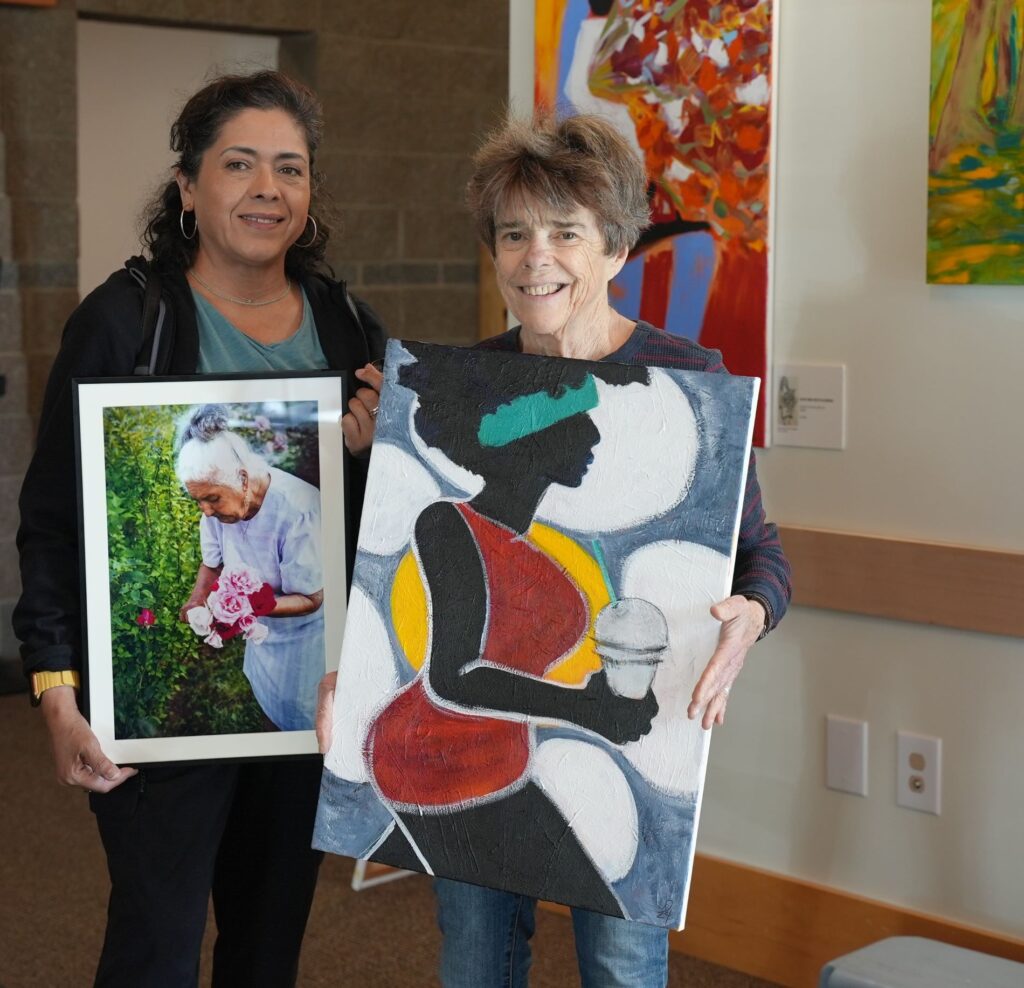 Local artists Sylvi Estrella and Laurie Davenport post with their respective art pieces for an upcoming Lakewood Arist Reception event. Sylvia holds a photograph of a woman gathering roses and Laurie holds a painting of a woman in silhouette holding a coffee drink.