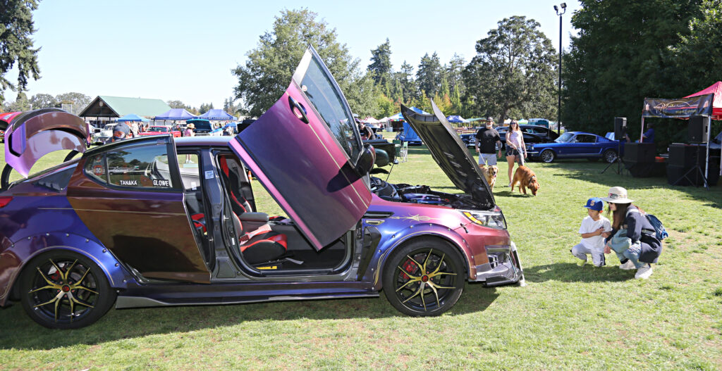 A woman and child crouch on the ground in front of a purple car on display with its doors open, front hood up and trunk open at a car show in Lakewood, WA.