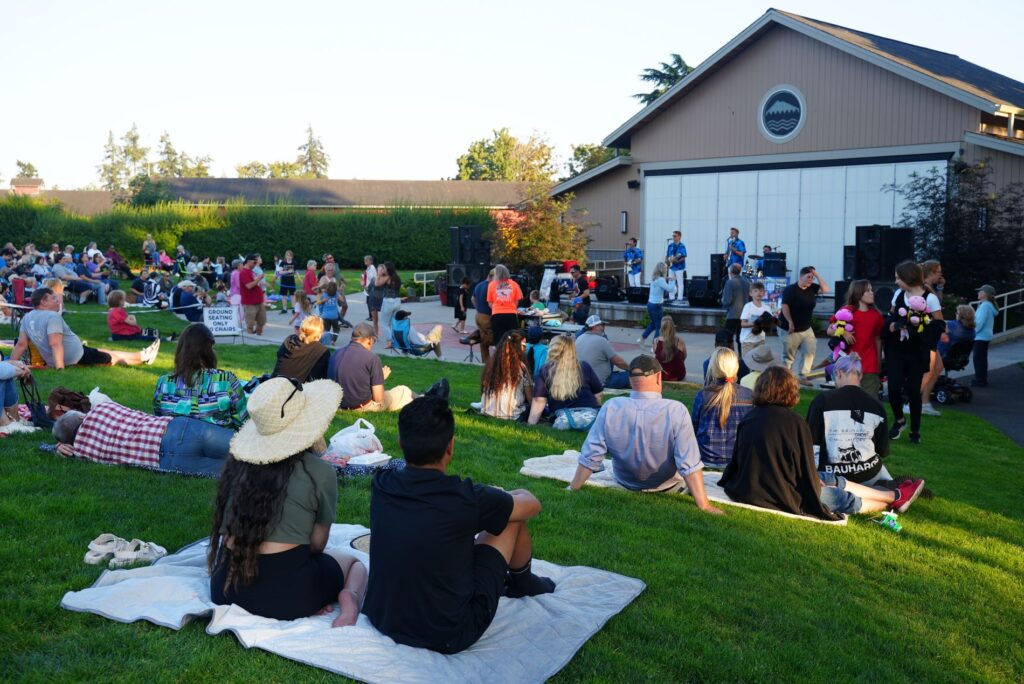 People sit on the grass at the Pavilion at Fort Steilacoom Park for a Summer Nights at the Pavilion summer concert in 2024. In the background a band plays on the stage and people are dancing.