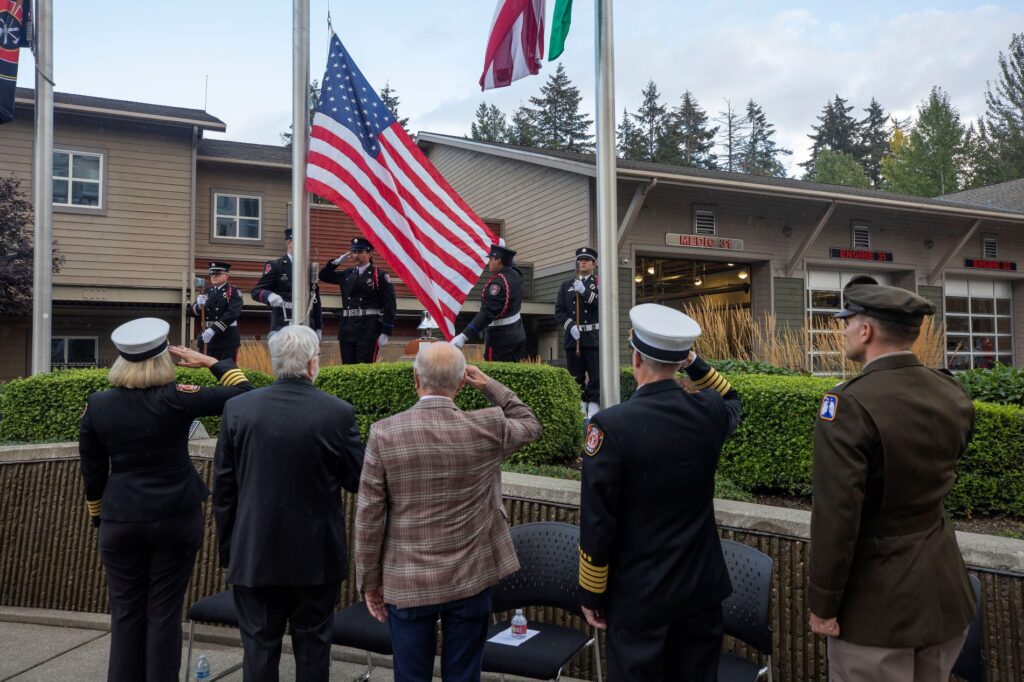 Firefighters and civilians stand with their backs to the camera facing the American Flag being risen up a flagpole in front of a fire station to commemorate 9/11 in 2024.