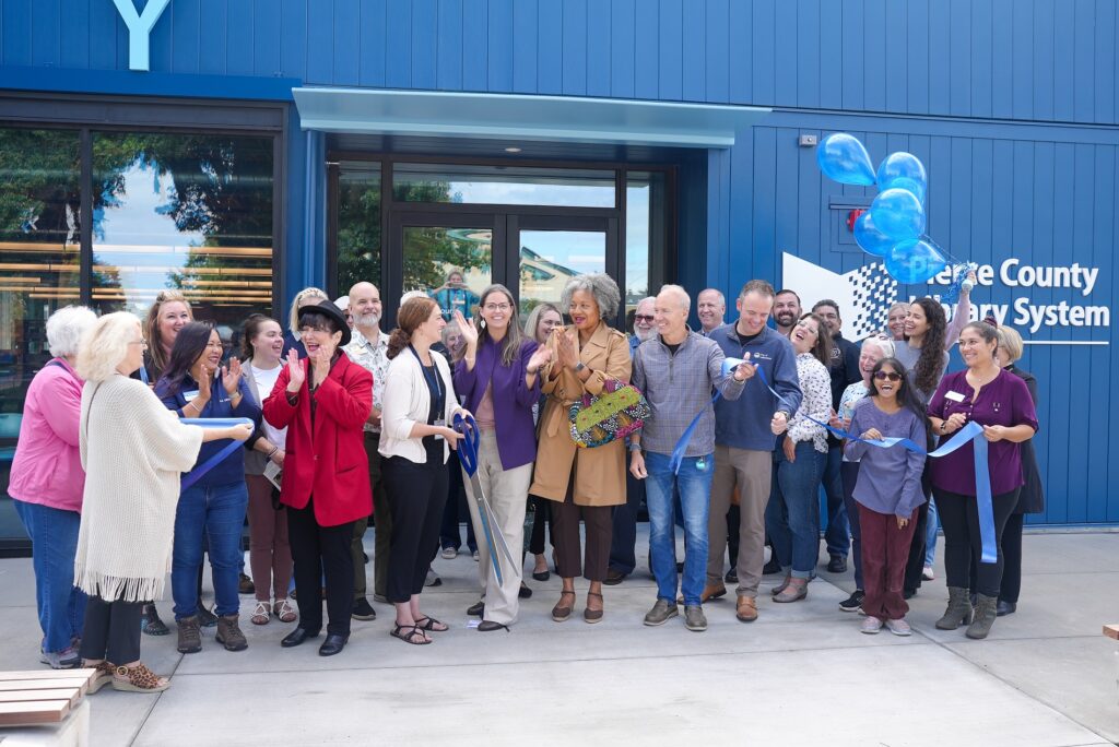 A group of people stand in front of the interim Lakewood library. They are cheering and just cut a blue ribbon to signal the library being open. A person holds a handful of blue balloons in the right corner of the frame.