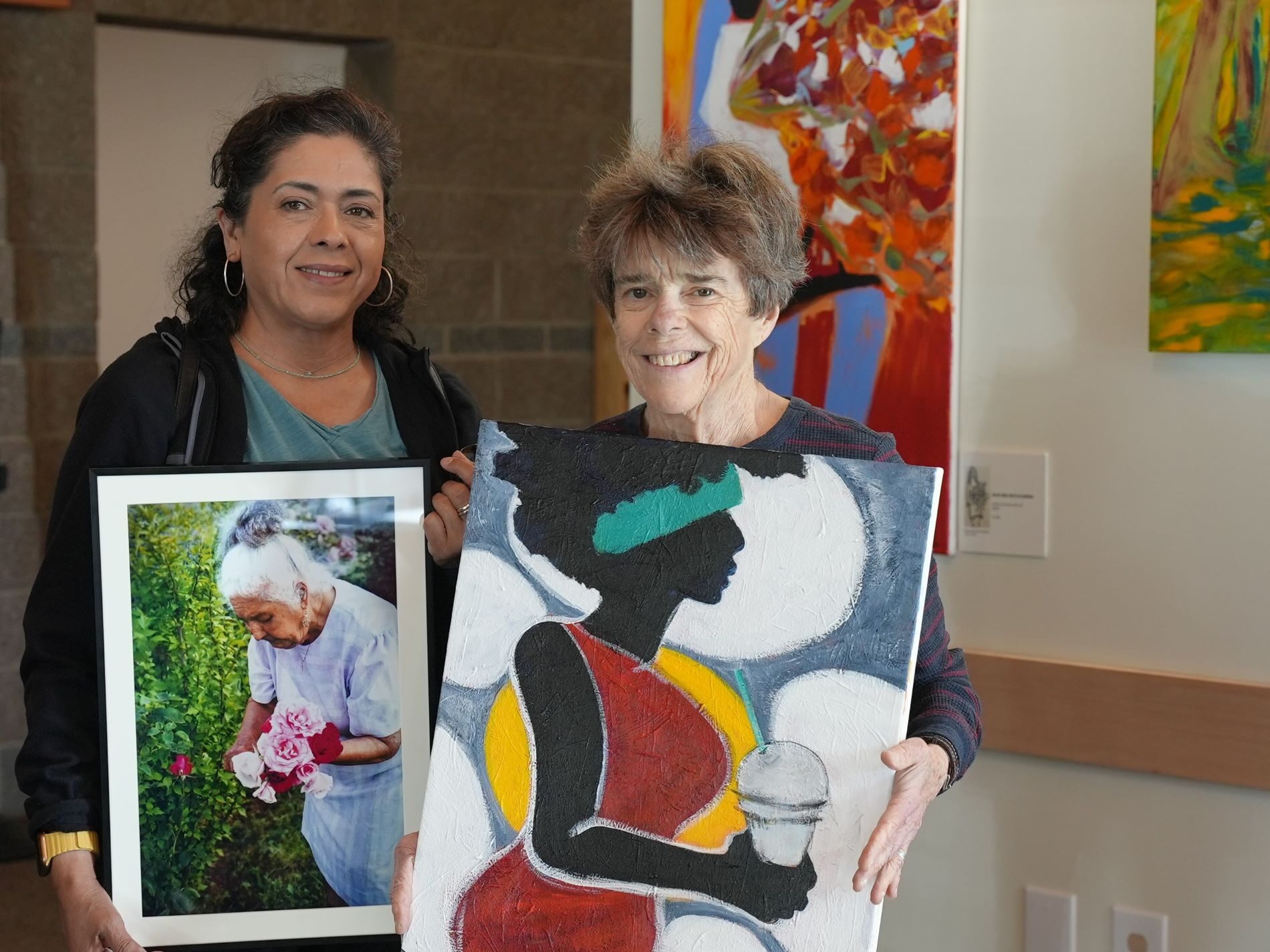 Local artists Sylvi Estrella and Laurie Davenport post with their respective art pieces for an upcoming Lakewood Arist Reception event. Sylvia holds a photograph of a woman gathering roses and Laurie holds a painting of a woman in silhouette holding a coffee drink.