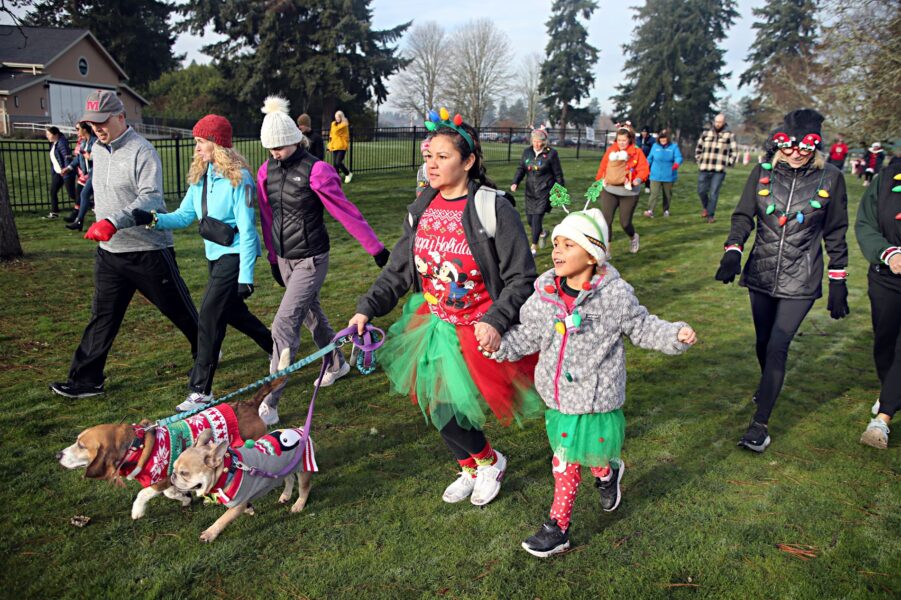 People dressed in holiday costumes participate in the 2023 Jingle Bell 5k walk/run at Fort Steilacoom. A woman walks two dogs on a leash dressed in costumes.