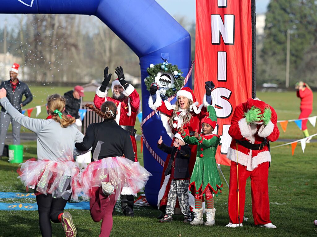 People cross the finish line at the 2023 Jingle Bell Fun Run in Fort Steilacoom Park. They are dressed in holiday costumes and a Santa, Mrs. Claus and Grinch are on the sideline cheering them on.