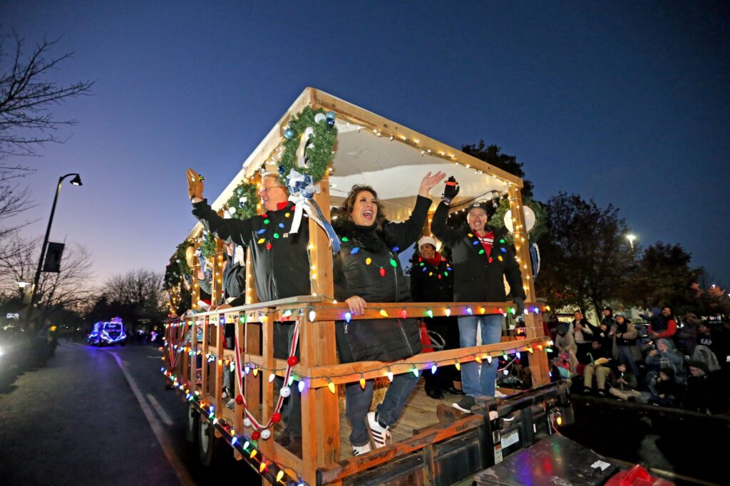 Members of the Lakewood City Council ride in a parade float lit with Christmas lights and decorated with green wreaths and wave to the crowd.