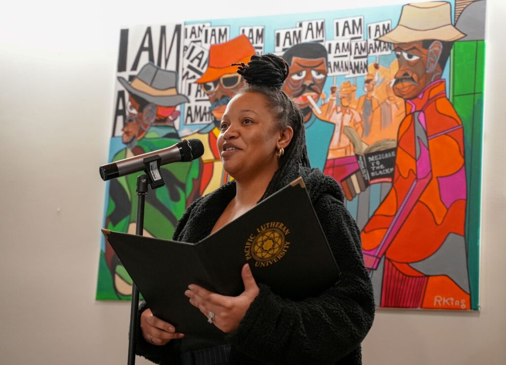 Spoken word artist Shaidaja Wilson-Dunham reads an original piece she wrote for the Beloved Community event. She stands at a microphone with a colorful painting behind her.