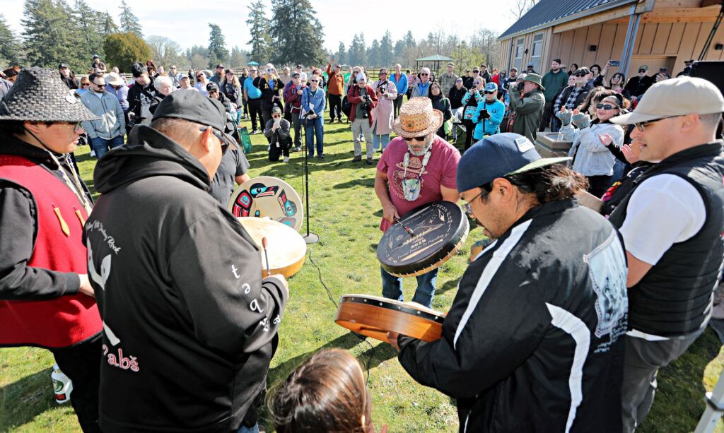 A group of men stand in a circle before a crowd playing traditional Native American handheld drums.