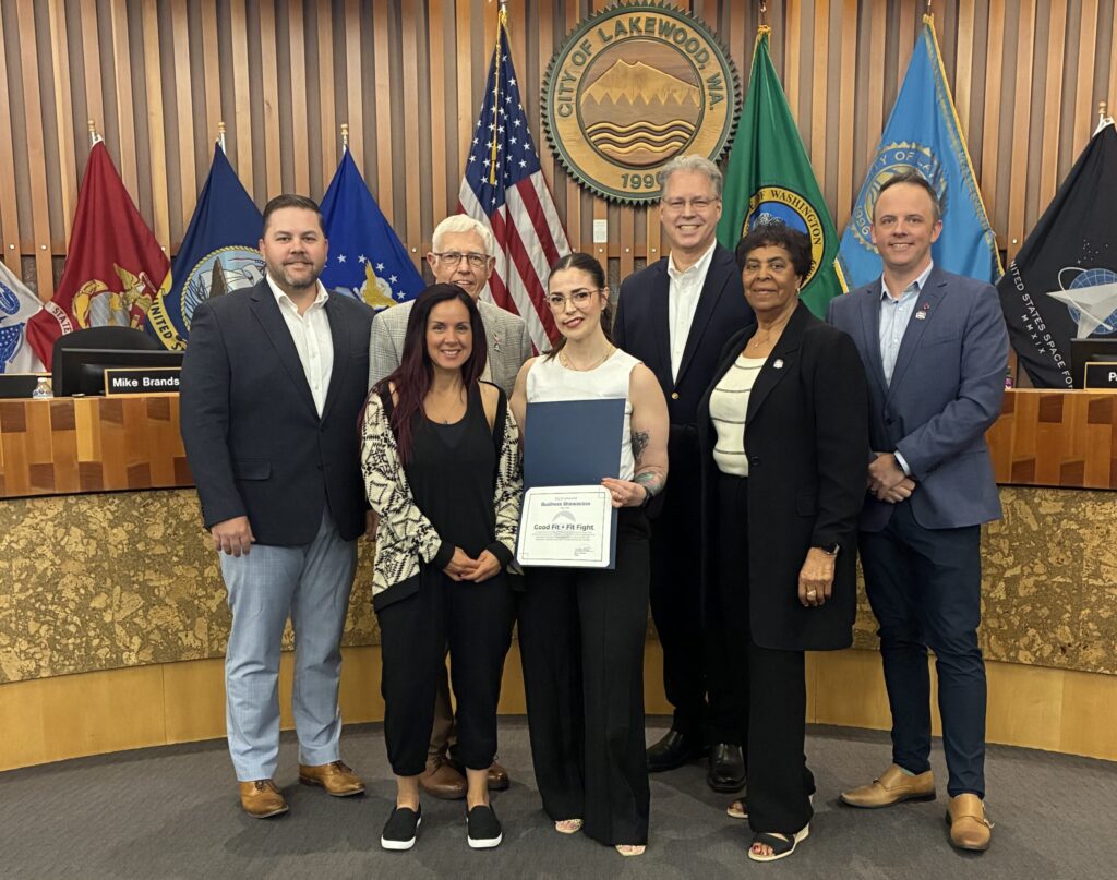 Members of the Lakewood City Council stand with business owners of Good Fit + Fit Fight, a local gym that was recognized as the May 2025 Business Showcase. They are in the Lakewood City Council chambers with flags in the background.