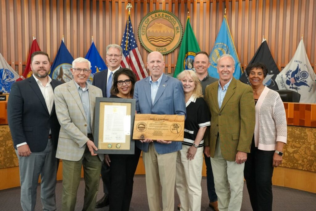 City Manager John J. Caulfield statnds in the middle of the Lakewood City Council in City Council Chambers holding a wooden plaque. The city seal and flags are lined up behind them.