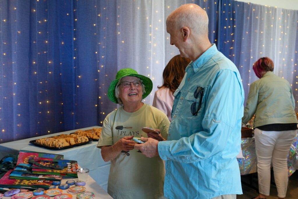 A couple look at each other and smile while holding Lakewood SWAG for volunteers at a volunteer recognition event. Behind them twinkle lights shine.