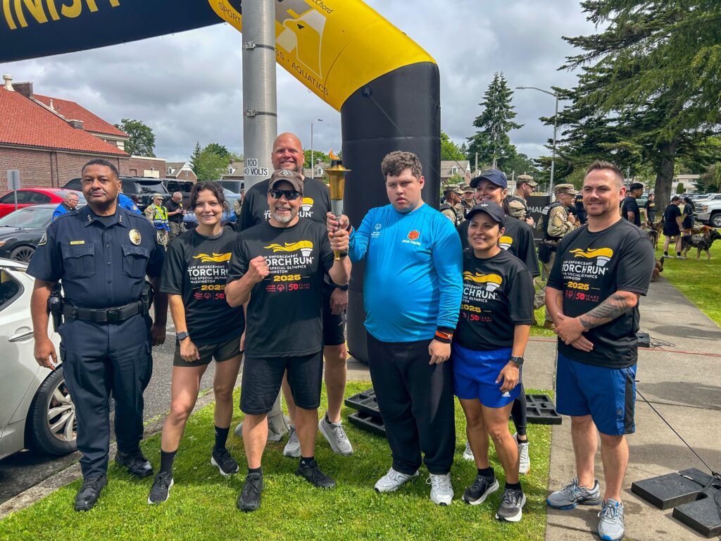 Police Chief Patrick D. Smith and members of the Lakewood Police Department stand with a participant of the 2025 Special Olympics Torch Run in front of a black and yellow inflatable arch. The participant is holding the torch and wearing a blue shirt.