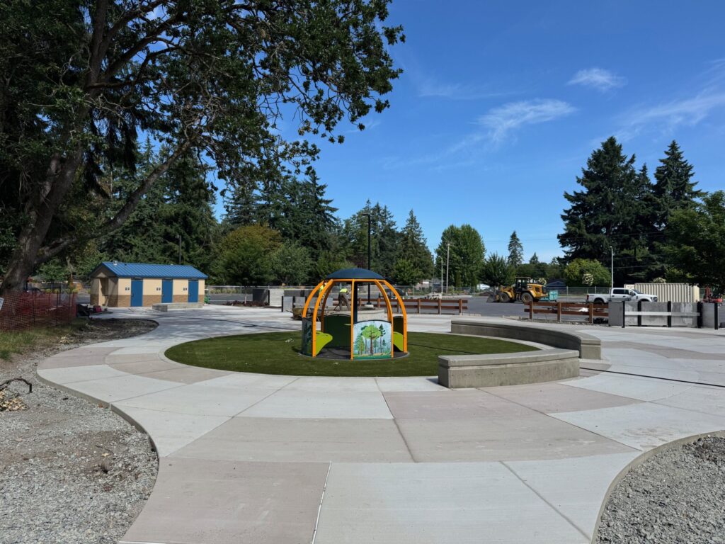A small play structure in a dome shape encircled by a patterned cement walkway and a bathroom in the distance at an outdoor park setting with a blue sky and large trees.