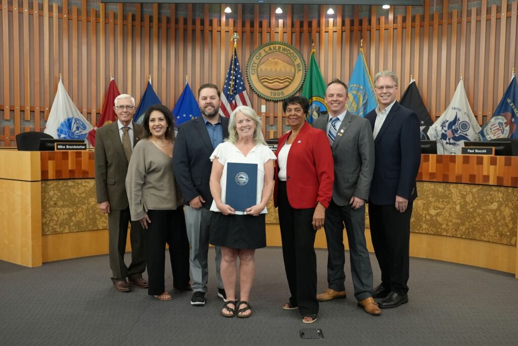 The Lakewood City Council stands at the front of the dais with retired Parks and Recreation director Mary Dodsworth who is holding a proclamation. Behind them are flags and the city seal for Lakewood.