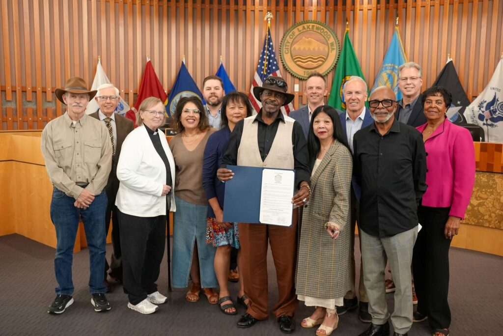 The Lakewood City Council poses with representatives from St. J-Js Episcopal Church who hold up a proclamation recognizing Juneteenth 2025. They are in Council Chambers with the city seal on the wall behind them and flags on display.