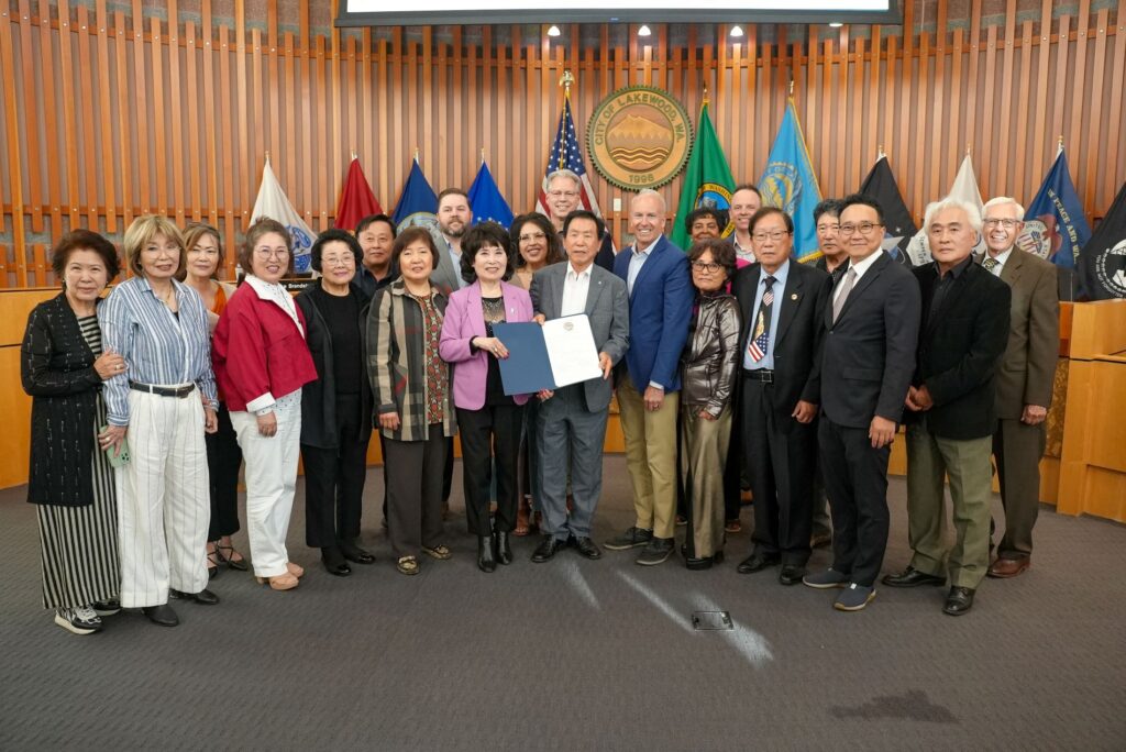 The Lakewood City Council poses with a large group of people from the Korean American community. Two people in the center of the group hold up a proclamation. They are in Council Chambers with the city seal on the wall behind them and flags on display.