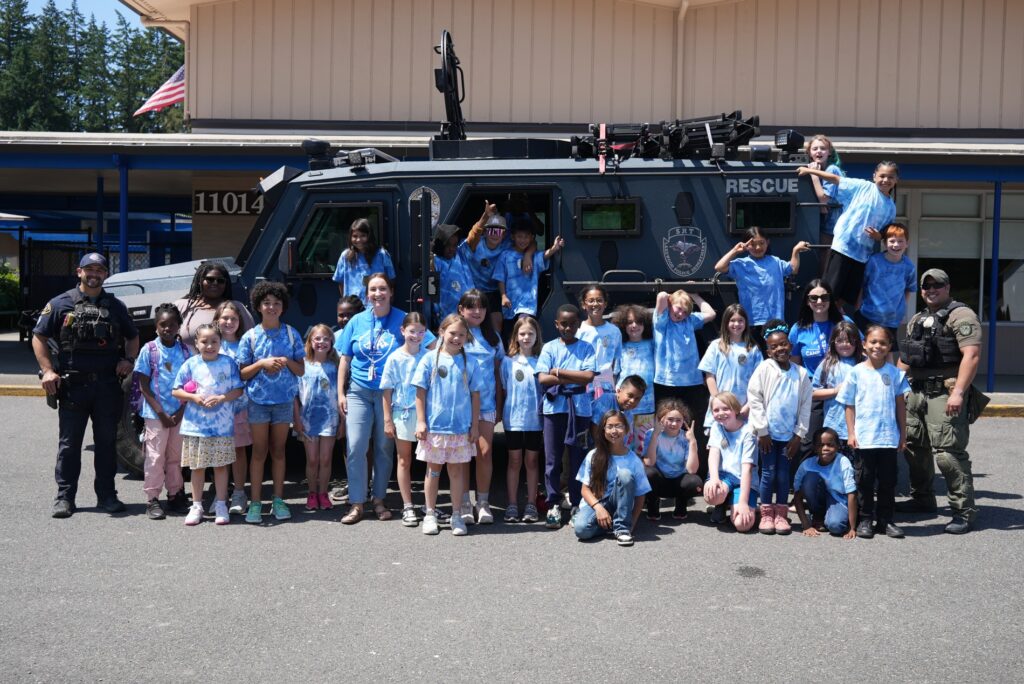 A group shot of students from Lake Louise Elementary School pose with two Lakewood Police officers in front of the department's SRT vehicle parked out front of the school. The students are in blue shirts, the officers in uniform. They are all smiling at the camera.