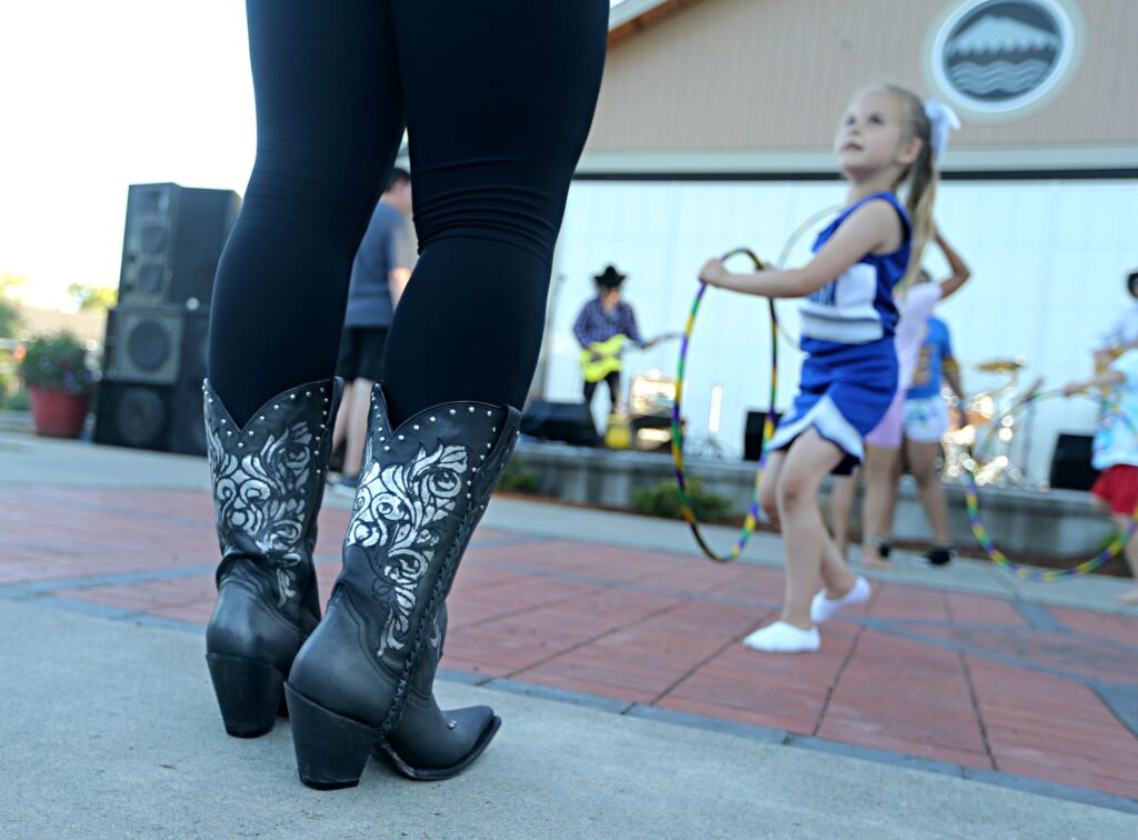 A close up of cowboy boots with a child dancing in the background and a band playing on an outdoor stage at a summer concert in Lakewood WA